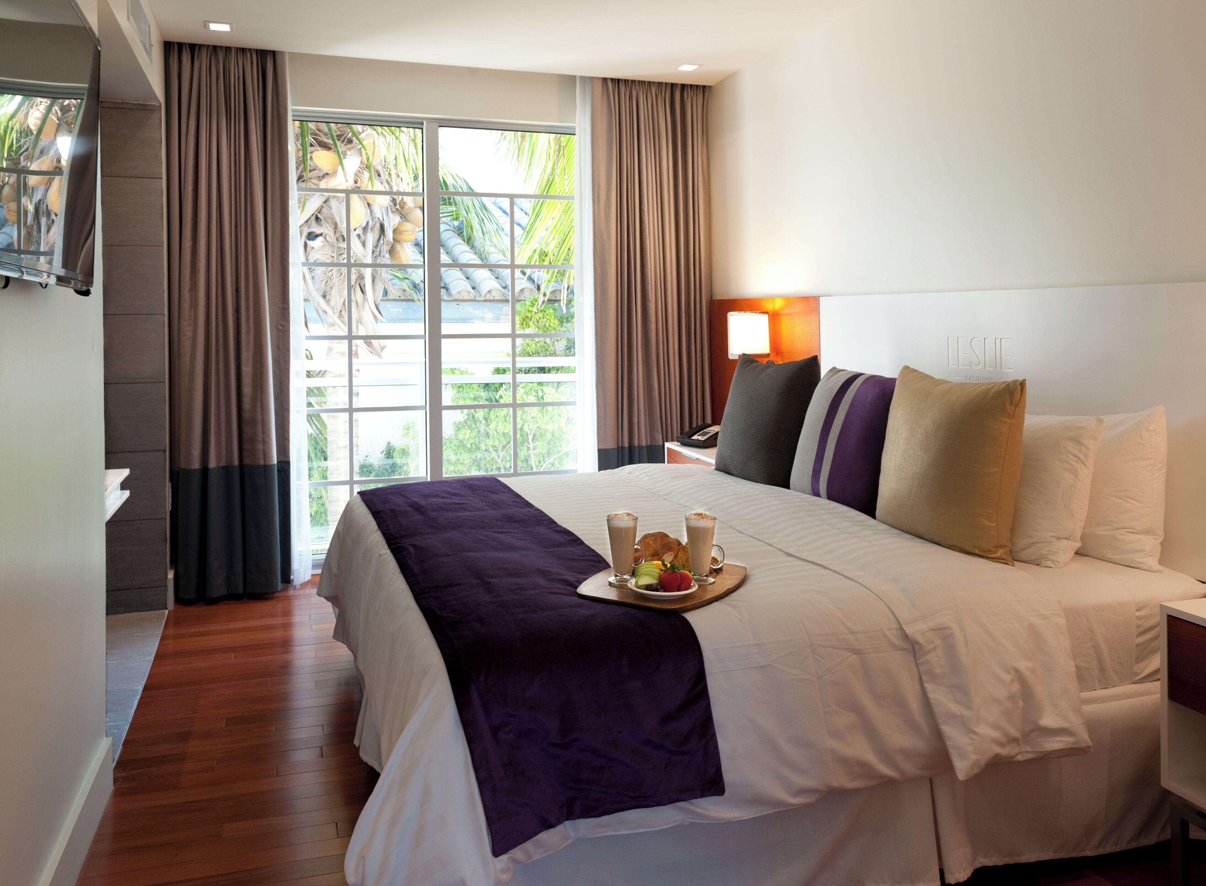 A hotel room with a bed, a bedside table, a lamp, and a view of palm trees through the window.