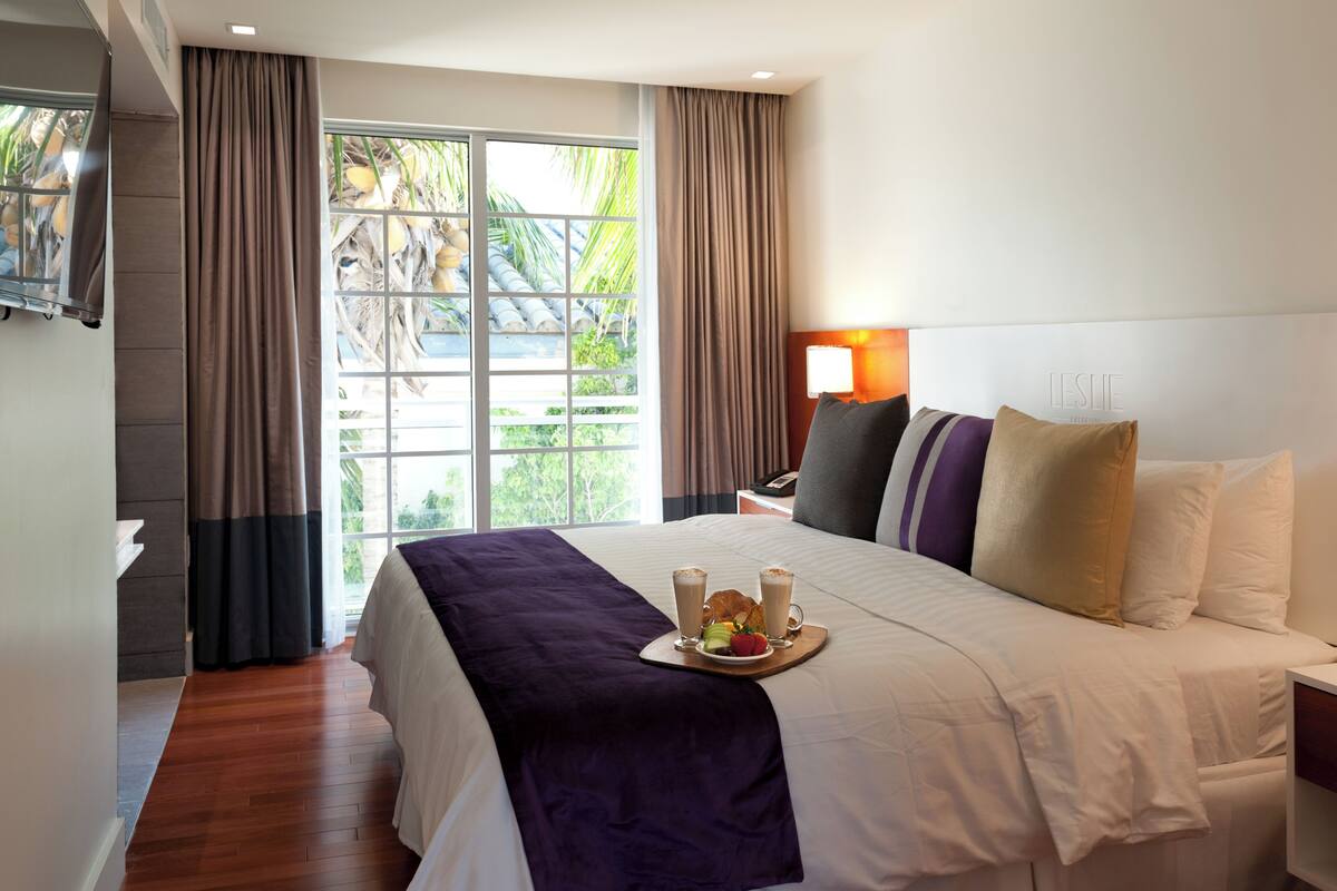 Una habitación de hotel con cama, mesita de noche, lámpara y vistas a palmeras a través de la ventana.