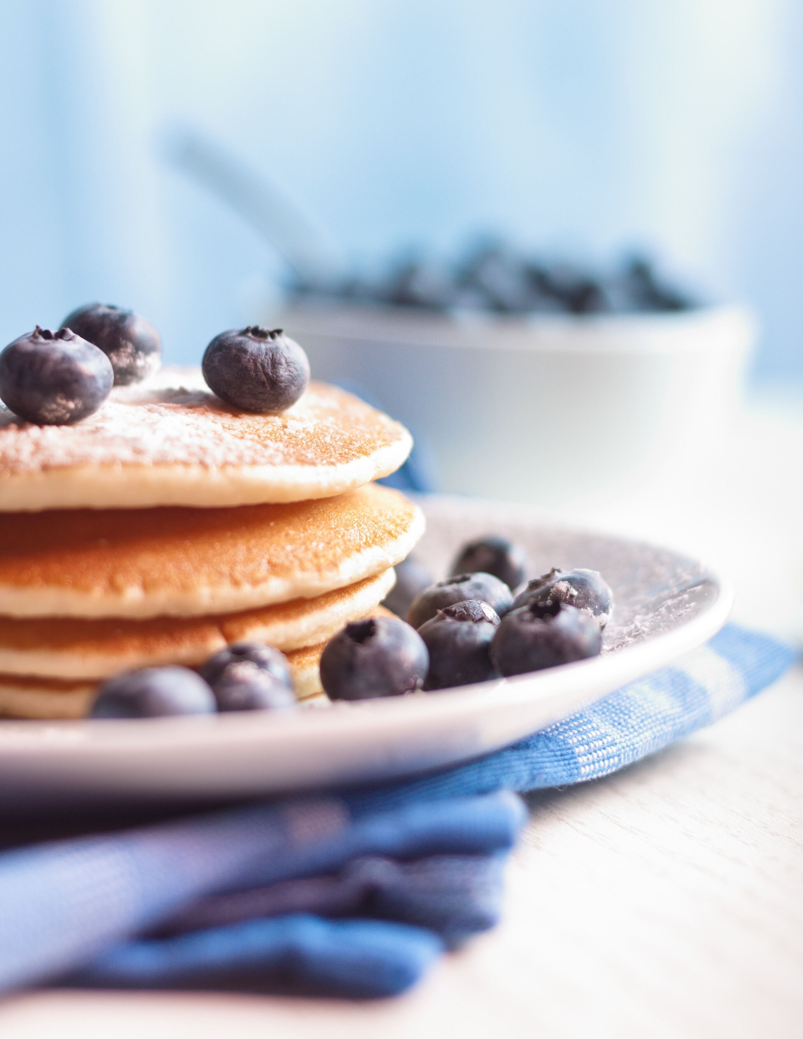 Une pile de crêpes garnies de myrtilles, présentées sur une assiette, avec un bol de myrtilles en arrière-plan.