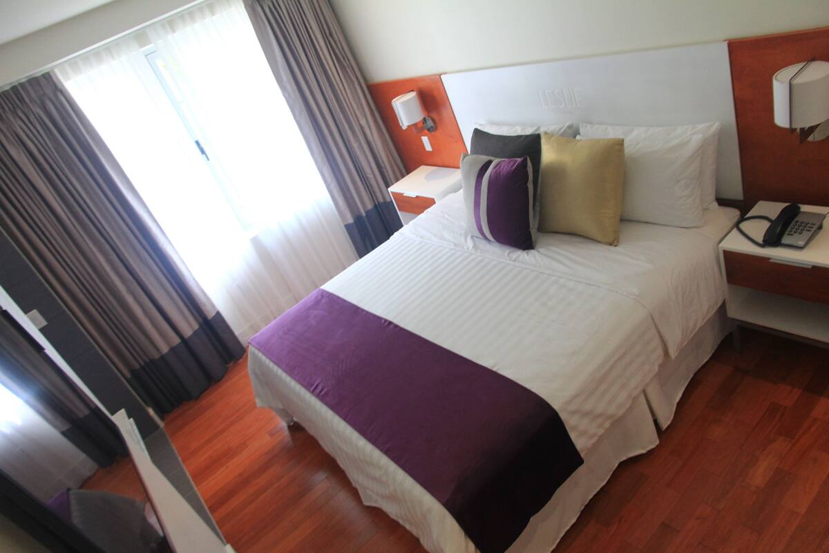 Una habitación de hotel con una cama grande, una mesita de noche con teléfono, una lámpara de pared y un ventanal con cortinas.