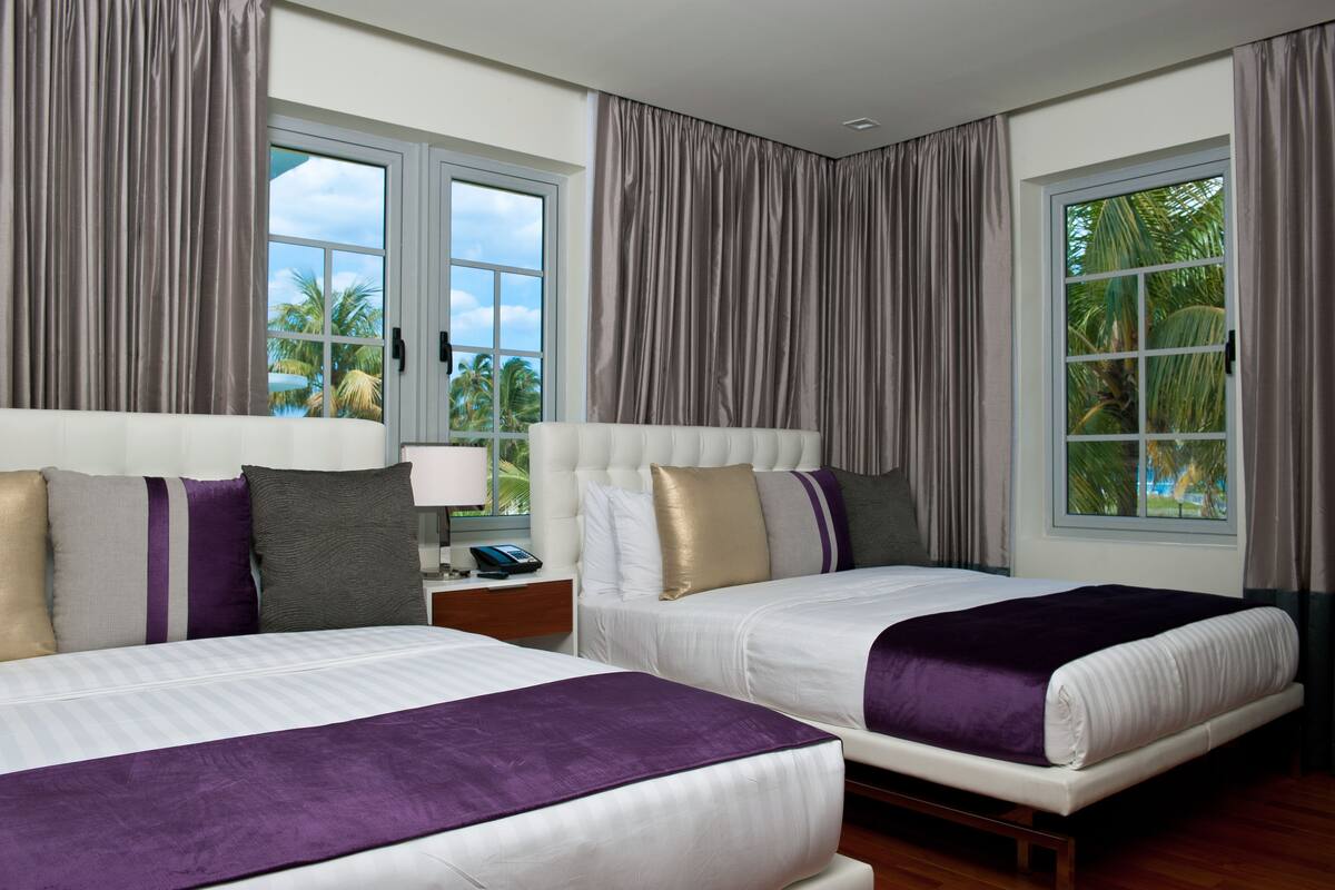 Habitación de hotel con dos camas, una mesita de noche, una lámpara y vistas a palmeras a través de las ventanas.