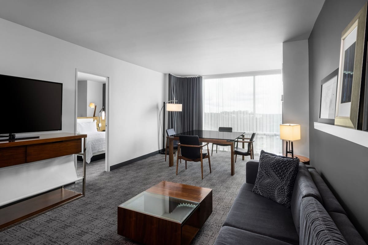 A modern hotel room with a flat-screen TV, a wooden desk, a sofa, a dining table, and a large window.