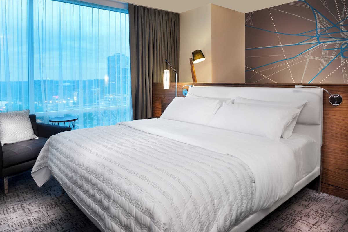 A hotel room with a large bed, a chair, a desk, and a view of a cityscape through large windows.