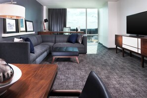 A modern hotel room with a grey sectional sofa, a small ottoman, a wooden coffee table, a flat-screen TV on a wooden stand, and a large window offering a city view.