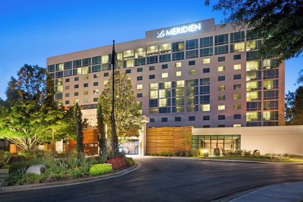 A modern hotel with a prominent sign reading "Le Meridien." It features a large glass facade, a well-maintained landscaped entrance, and a clear view of the building's exterior.
