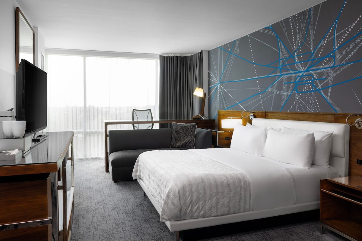 A modern hotel room with a large bed, a desk with a flat-screen TV, a chair, and a large window with curtains.
