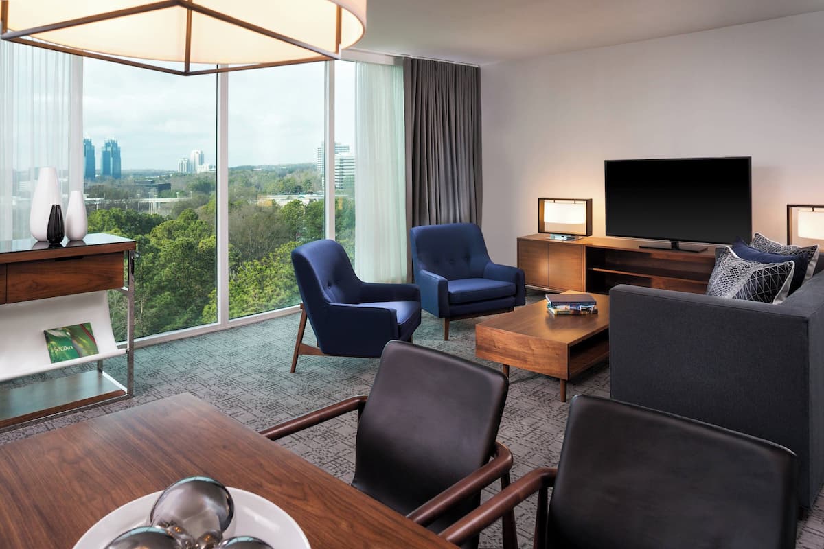A modern hotel room with a large window, a flat-screen TV, a sofa, two armchairs, a wooden coffee table, and a small side table.