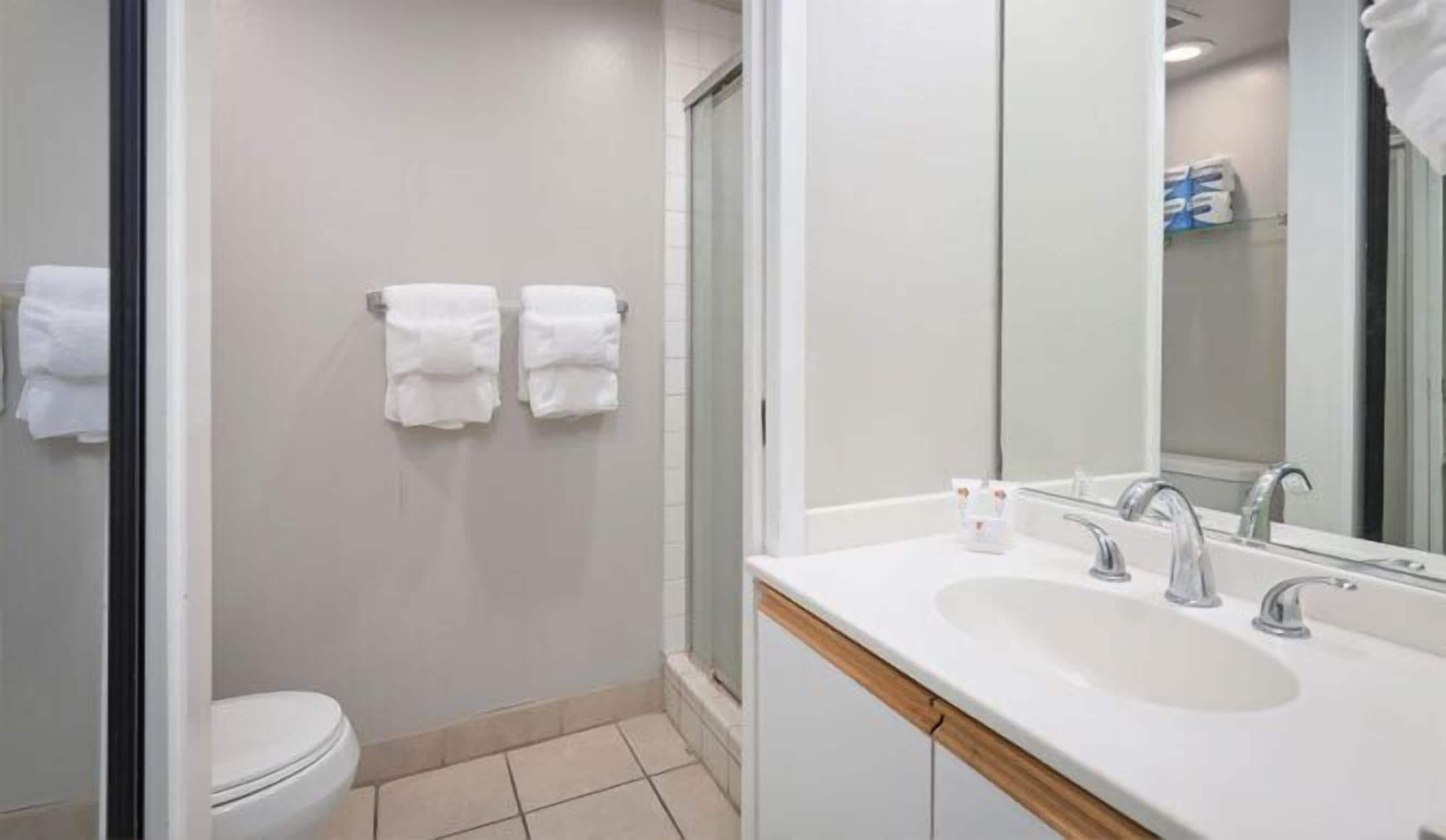 Un baño moderno con inodoro, dos toallas blancas colgadas en la pared, zona de ducha y lavamanos con espejo.