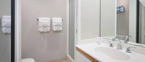 Un baño moderno con inodoro, dos toallas blancas colgadas en la pared, zona de ducha y lavamanos con espejo.