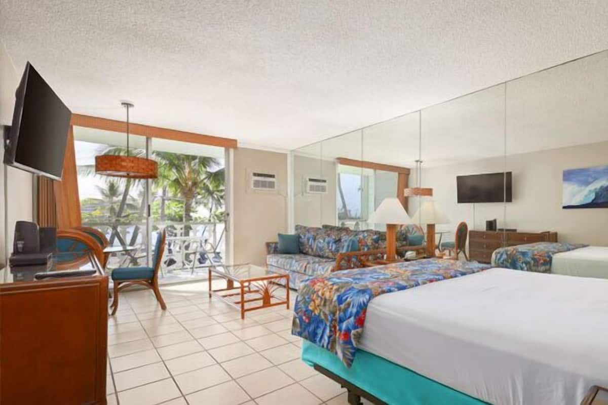 A hotel room with a bed, a desk, a chair, a TV, and a balcony with a view of palm trees.