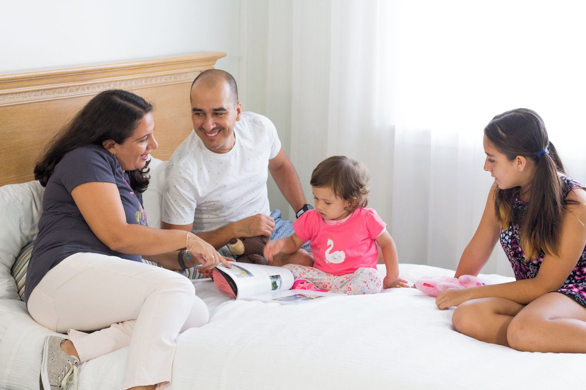 A family of four in a hotel room with a bed, a bedside table, and a window with curtains.