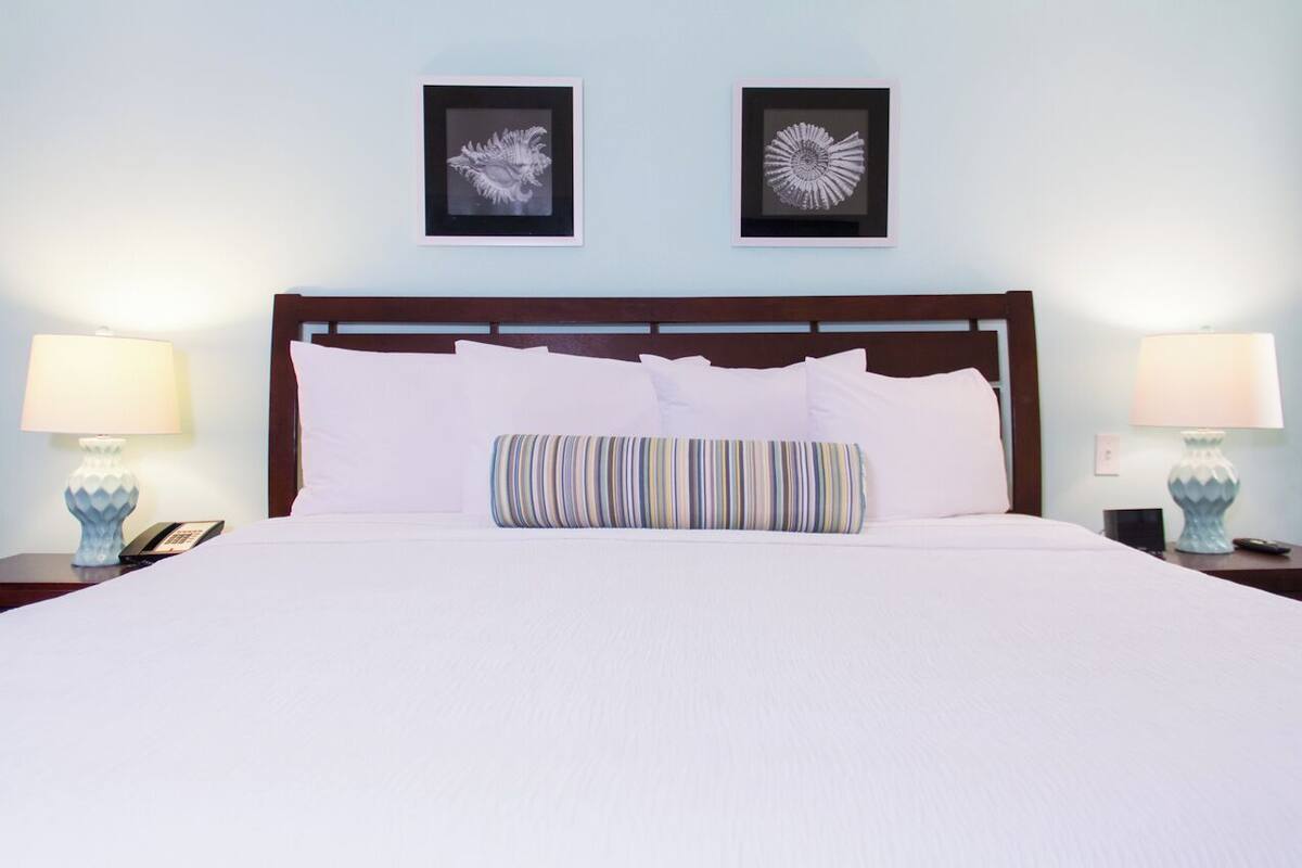 A neatly made bed with white bedding and a striped pillow, flanked by two bedside tables with lamps and a phone.