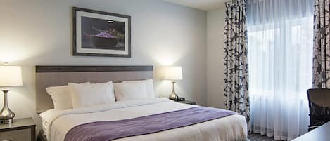 A hotel room with a bed, two bedside lamps, a desk, a chair, a framed picture on the wall, and a window with curtains.