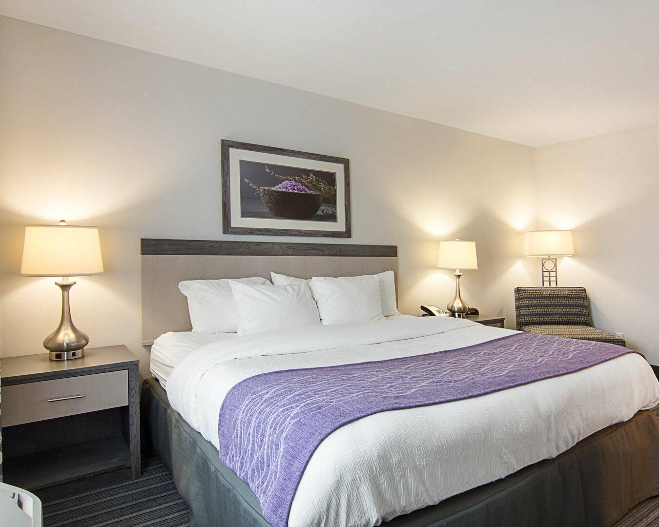 Une chambre d’hôtel avec un lit, deux tables de chevet, une petite table, une lampe, un tableau encadré au mur et un plaid à motifs.