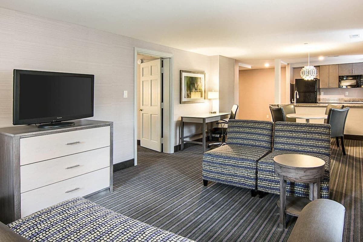 A hotel room with a flat-screen TV, a sofa, a dining table with chairs, a small kitchen area, and a door leading to another room.