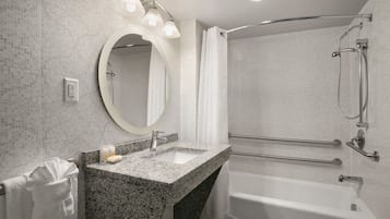 A modern bathroom with a granite sink, a round mirror, a shower with a handheld showerhead, and a grab bar.