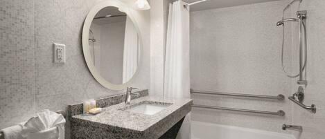 A modern bathroom with a granite sink, a round mirror, a shower with a handheld showerhead, and a grab bar.