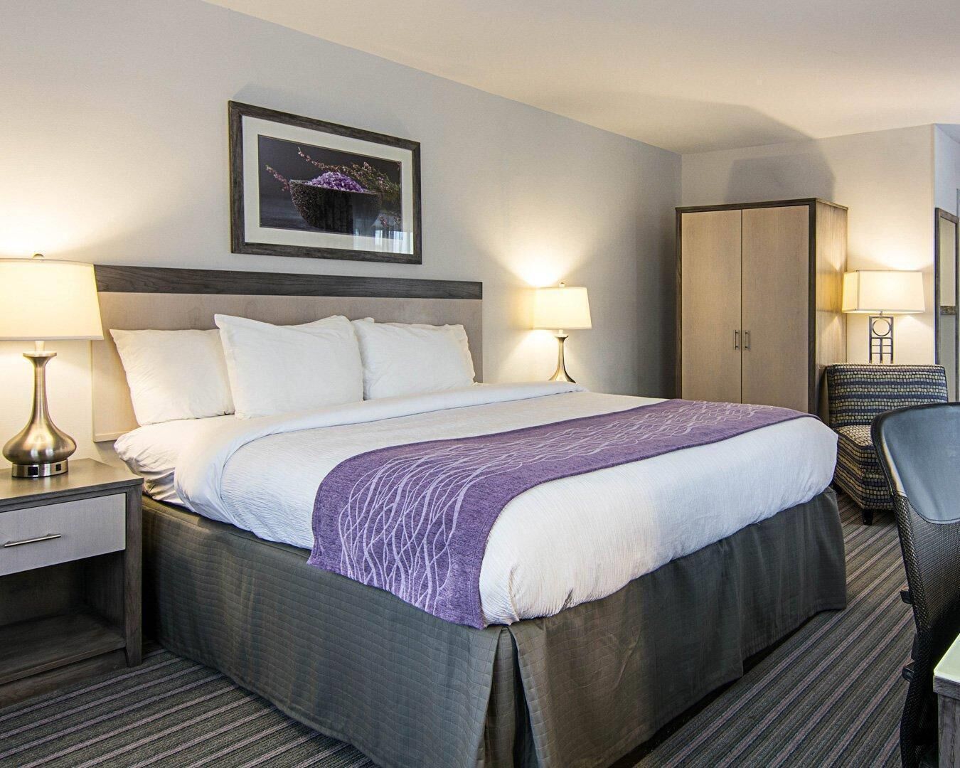 A hotel room with a bed, two bedside lamps, a chair, a wardrobe, and a framed picture on the wall.