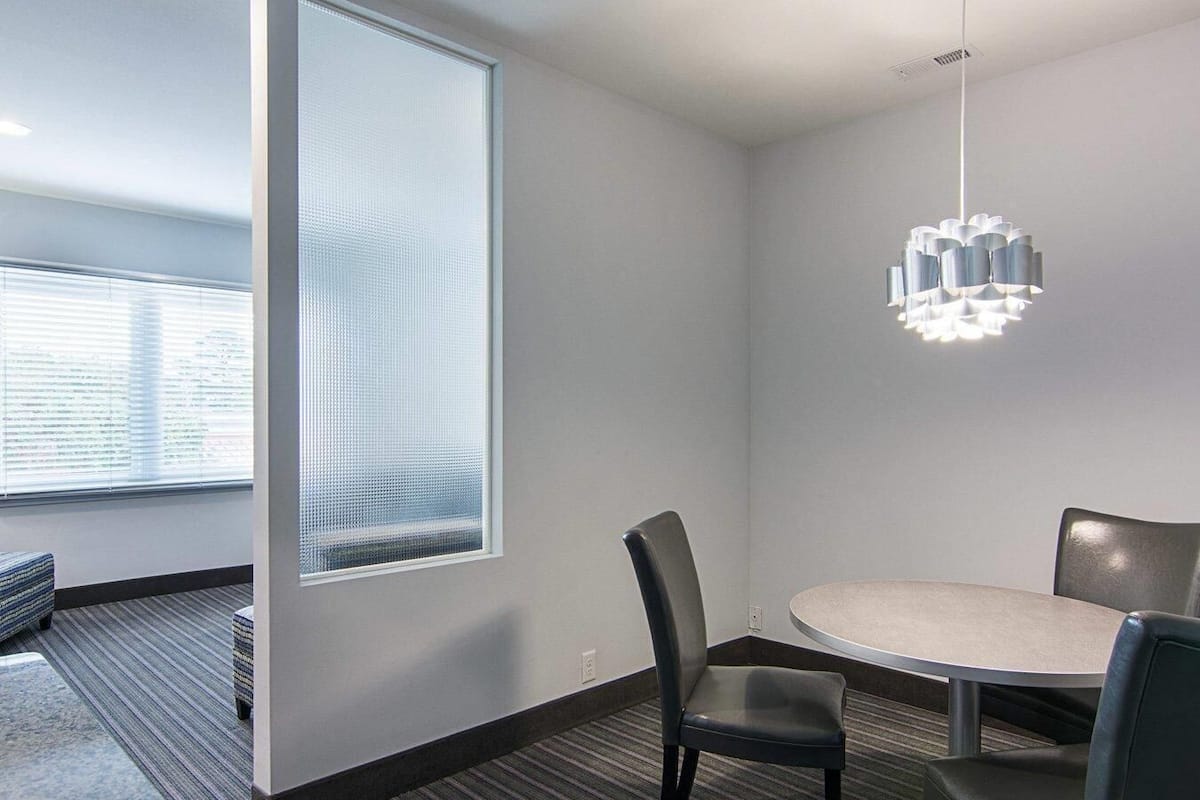 A modern meeting room with a round table, two chairs, a hanging light fixture, and a large window with blinds.