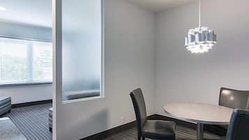 A modern meeting room with a round table, two chairs, a hanging light fixture, and a large window with blinds.