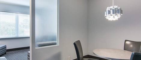 A modern meeting room with a round table, two chairs, a hanging light fixture, and a large window with blinds.