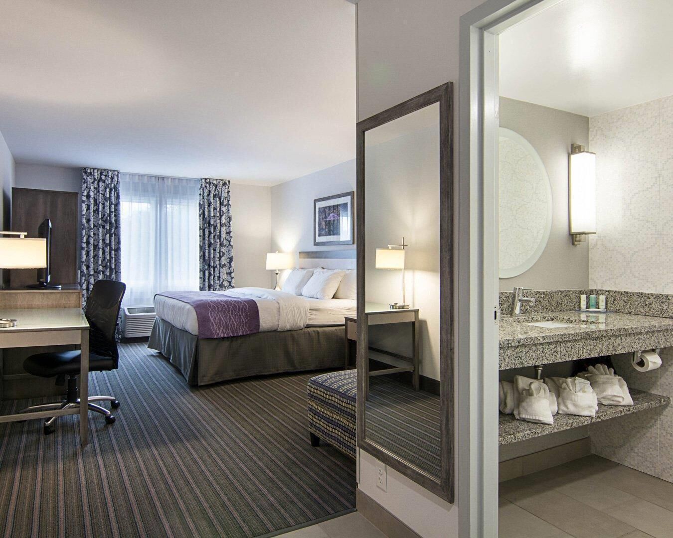 A hotel room with a bed, desk, chair, and a bathroom with a sink and mirror.