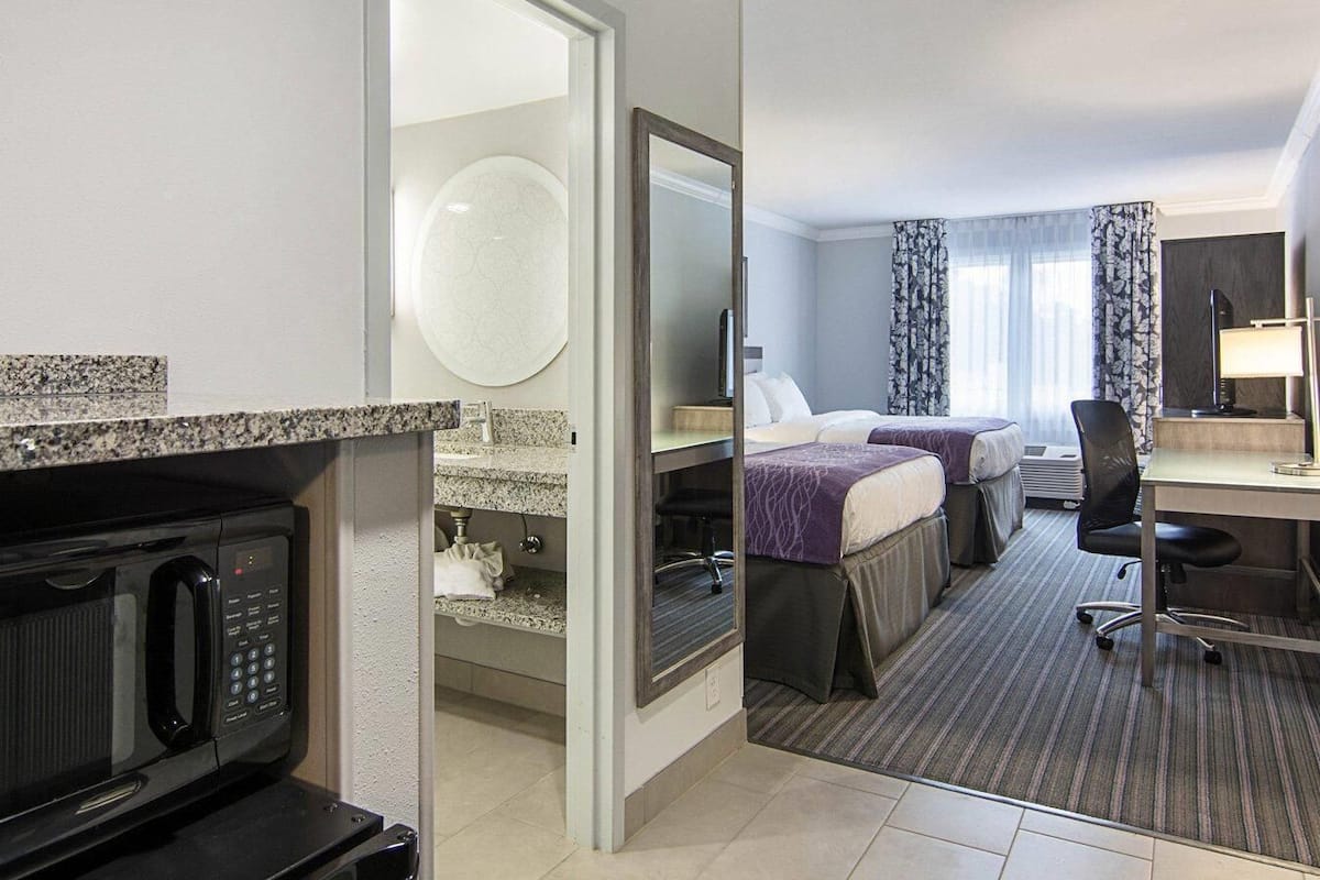 A hotel room with a kitchenette, two beds, a desk, and a large mirror.