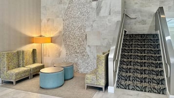 Eine Lobby mit einer Couch, zwei runden Tischen, einer Lampe, einer strukturierten Wand und einer gemusterten Treppe.