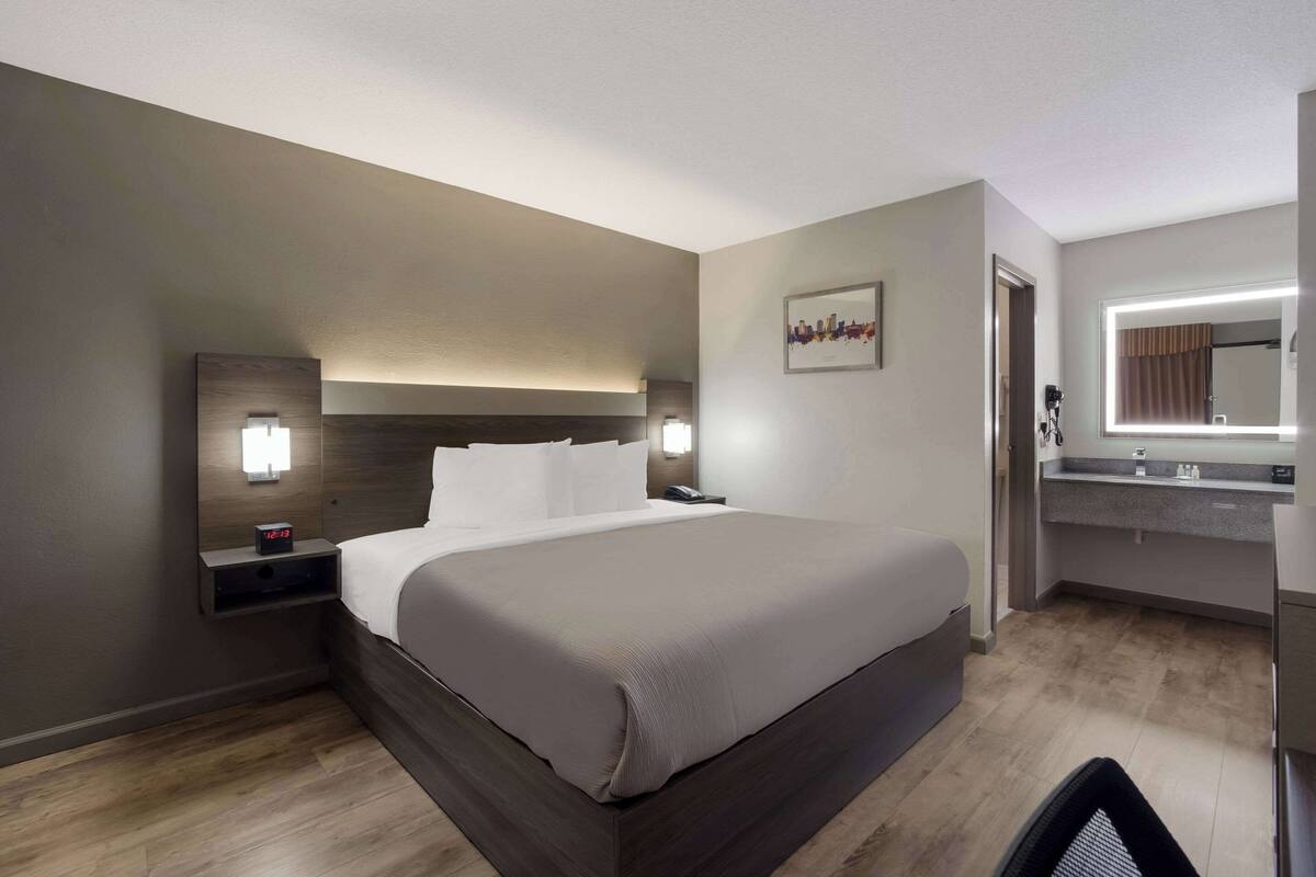 A modern hotel room with a large bed, bedside tables with lamps, a built-in desk, a mirror, and a bathroom with a vanity.