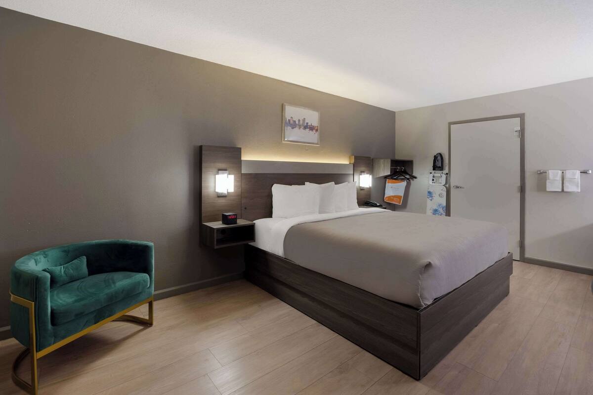 A hotel room with a large bed, a green armchair, bedside tables with lamps, and a cityscape artwork on the wall.