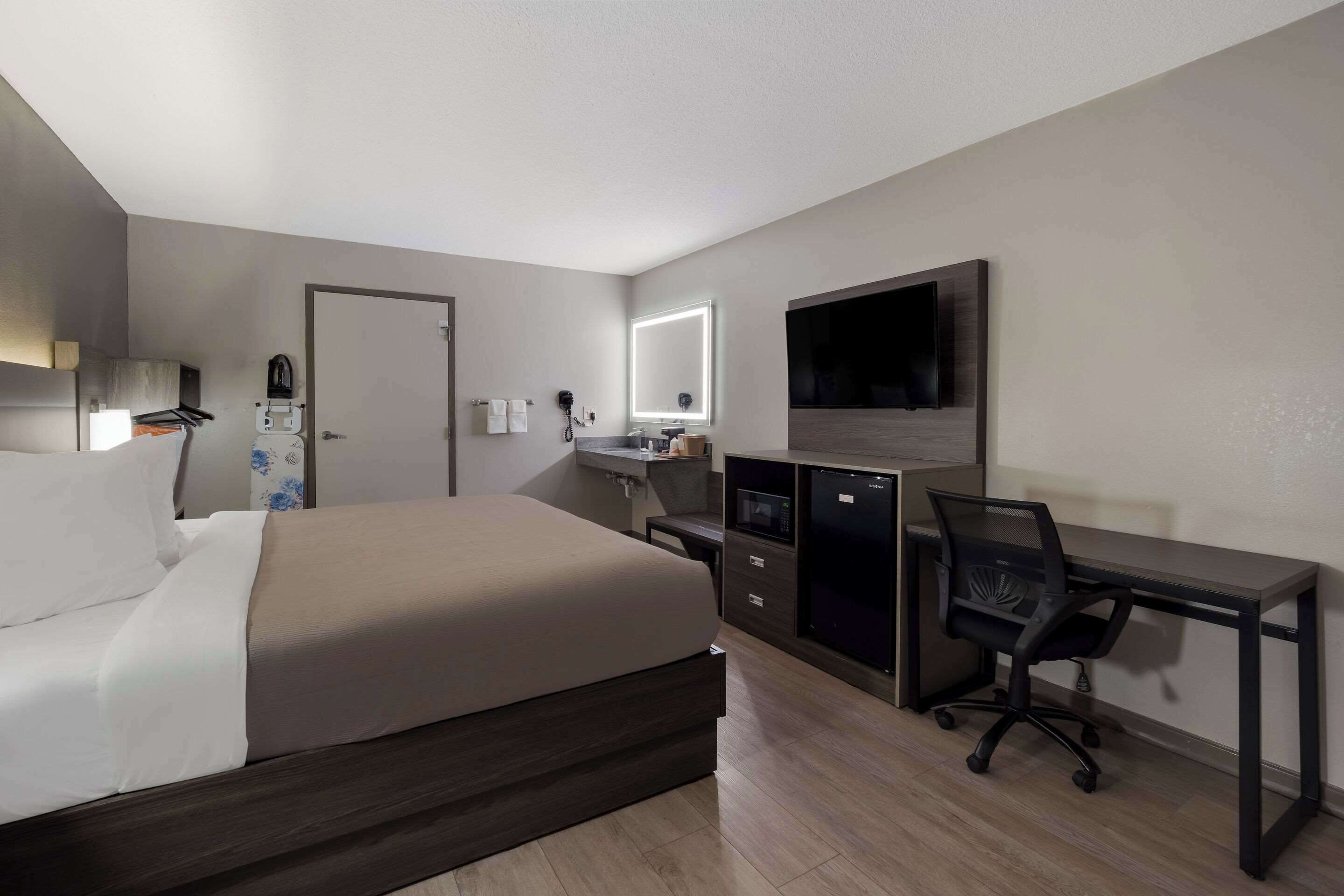 A hotel room with a bed, a desk with a chair, a TV, a mirror, and a small kitchenette.