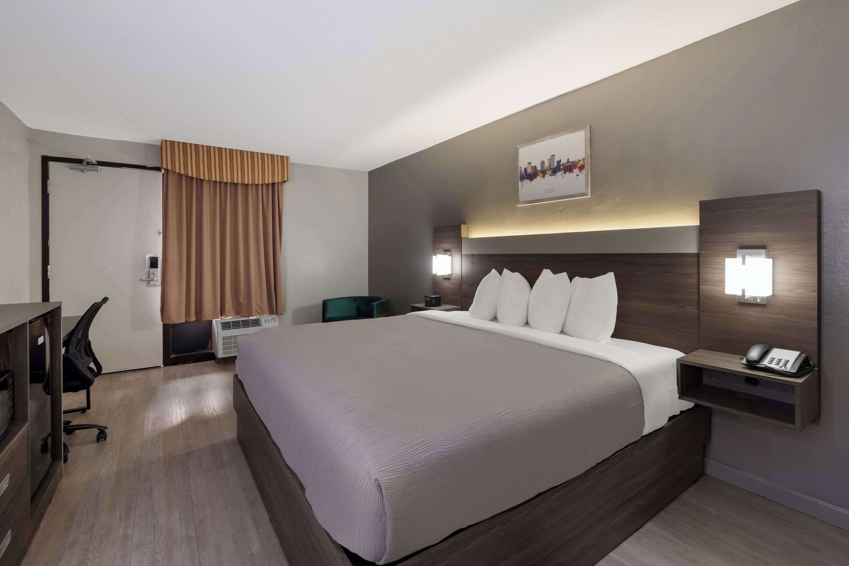 Une chambre d’hôtel moderne avec un grand lit, un bureau et une chaise. On y trouve une table de chevet avec un téléphone et une lampe.