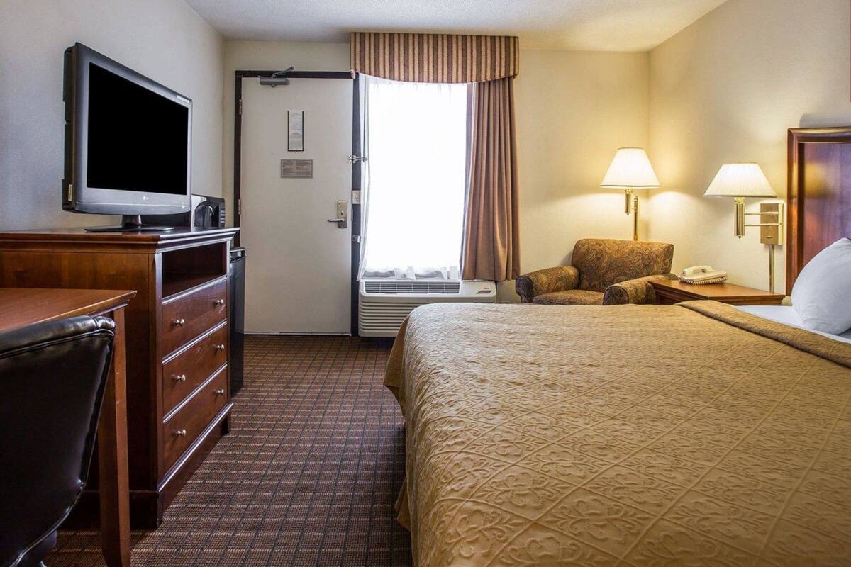 A hotel room with a bed, a television, a desk with drawers, a chair, a lamp, and a window with curtains.