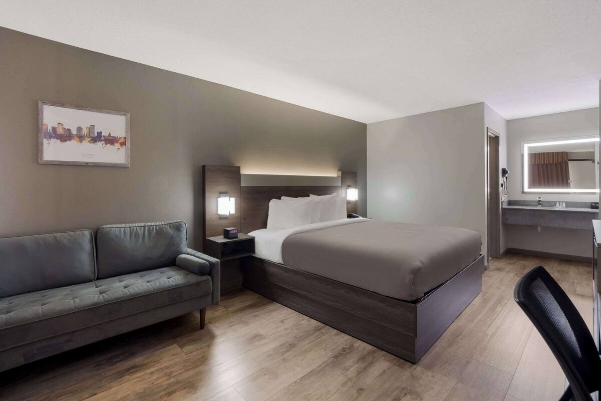 A hotel room with a bed, a grey sofa, a nightstand, a desk, and a mirror.