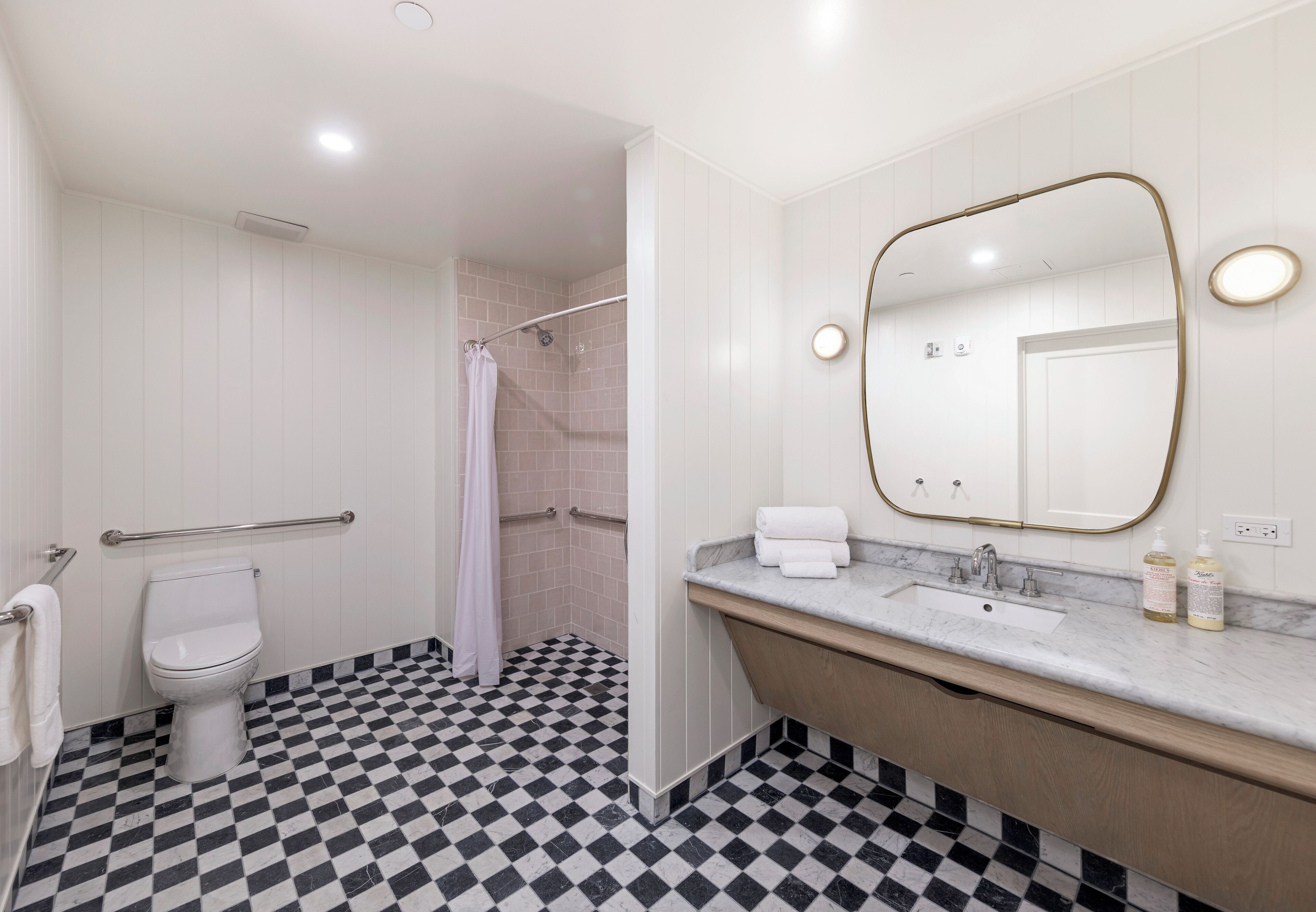 A bathroom with a checkered floor, a white toilet, a shower, a sink with a marble countertop, and a large mirror with a gold frame.