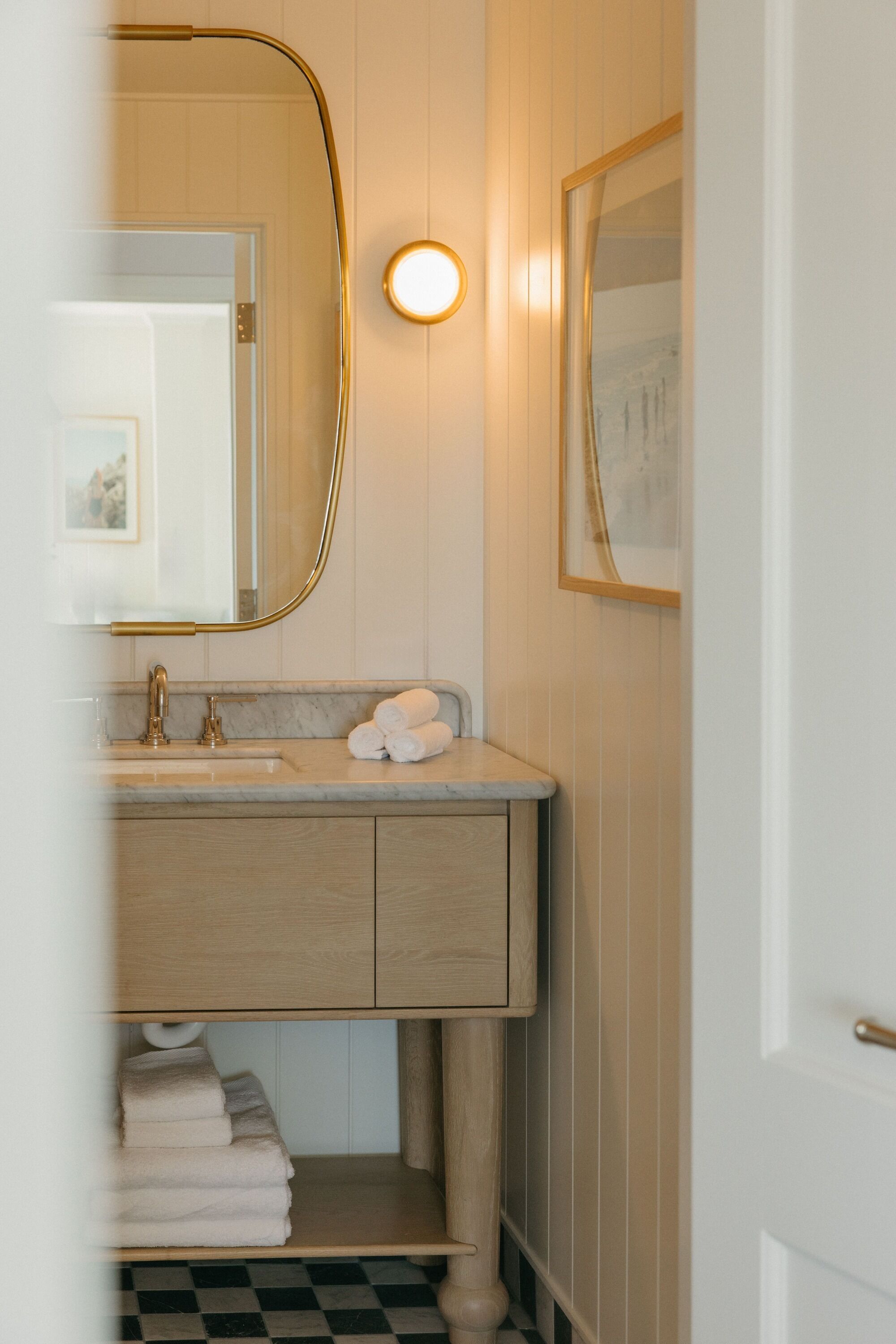 Une salle de bain avec un meuble-lavabo en bois, un miroir avec un cadre doré, un applique murale ronde et un tableau encadré accroché au mur.