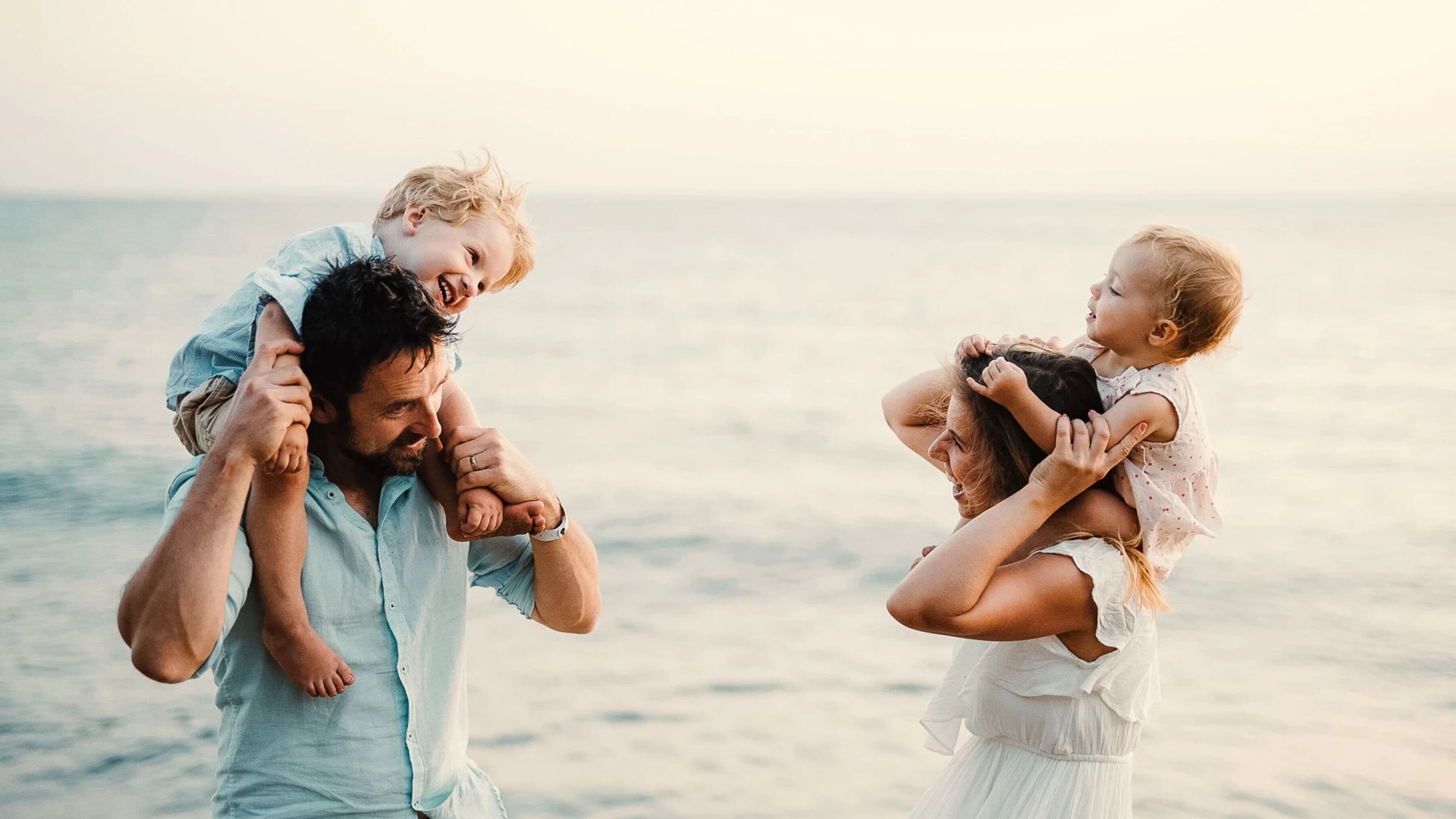 Een gezin van drie op het strand, waarbij de vader twee kinderen op zijn schouders draagt.