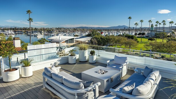 A modern outdoor deck with a sofa, chairs, and a table, overlooking a marina with boats and palm trees.