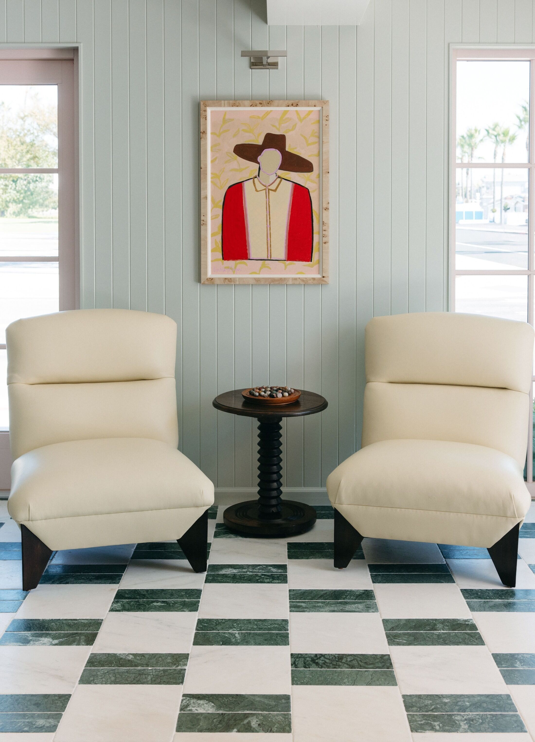A room with two cream-colored armchairs, a small round table with a bowl of fruit, and a framed artwork on the wall.