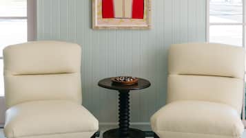 A room with two cream-colored armchairs, a small round table with a bowl of fruit, and a framed artwork on the wall.
