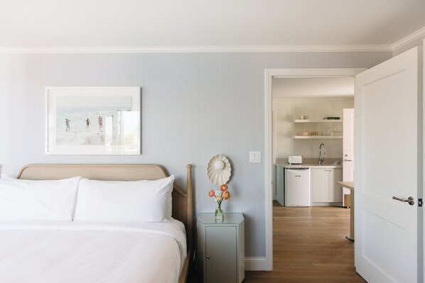 Un cuarto de hotel con una cama, una mesita de noche con un florero y un cuadro en la pared, además de vistas hacia la zona de la cocina.
