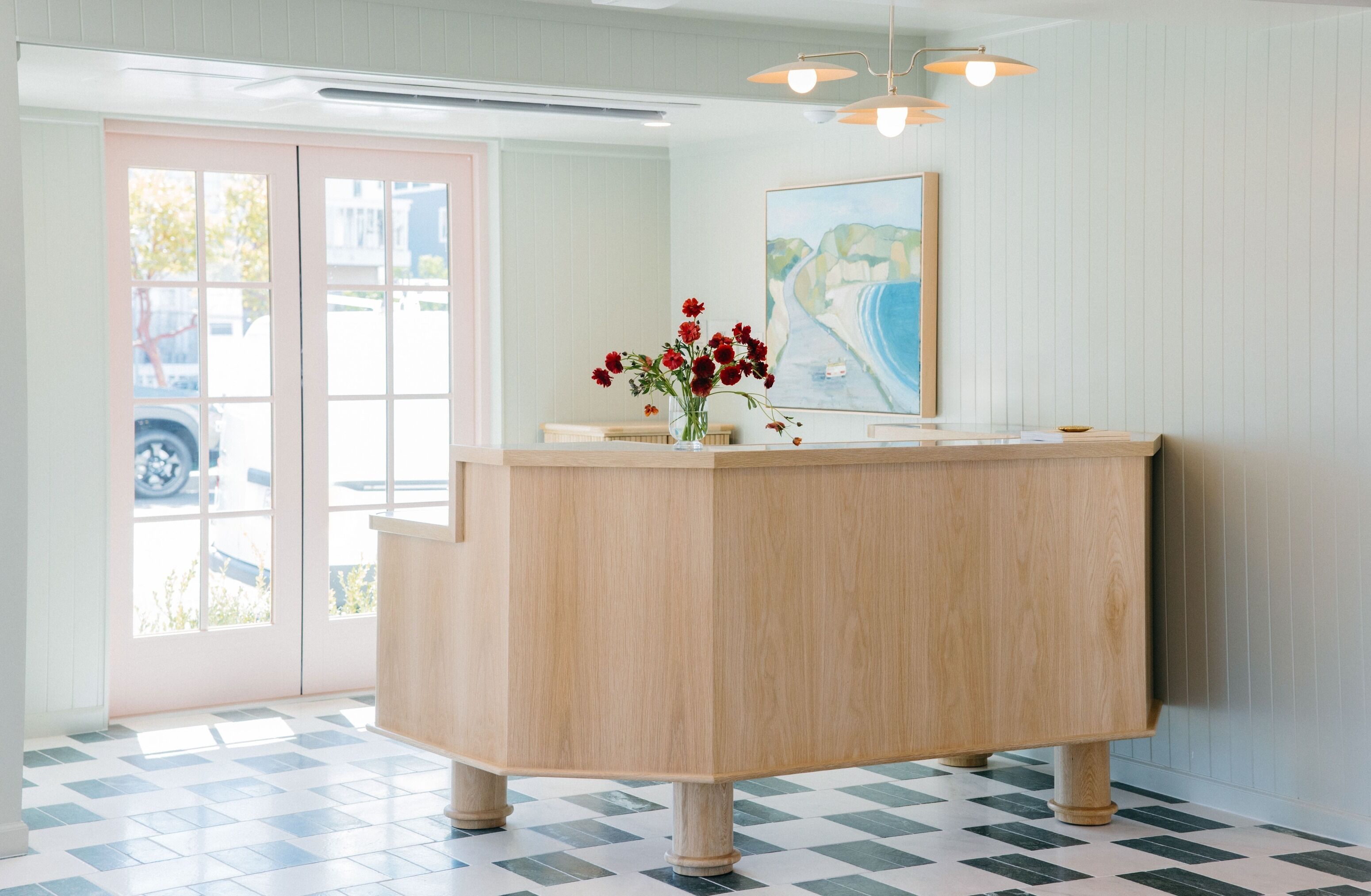A reception area with a wooden reception desk, a vase with red flowers, a painting on the wall, and a checkered floor.