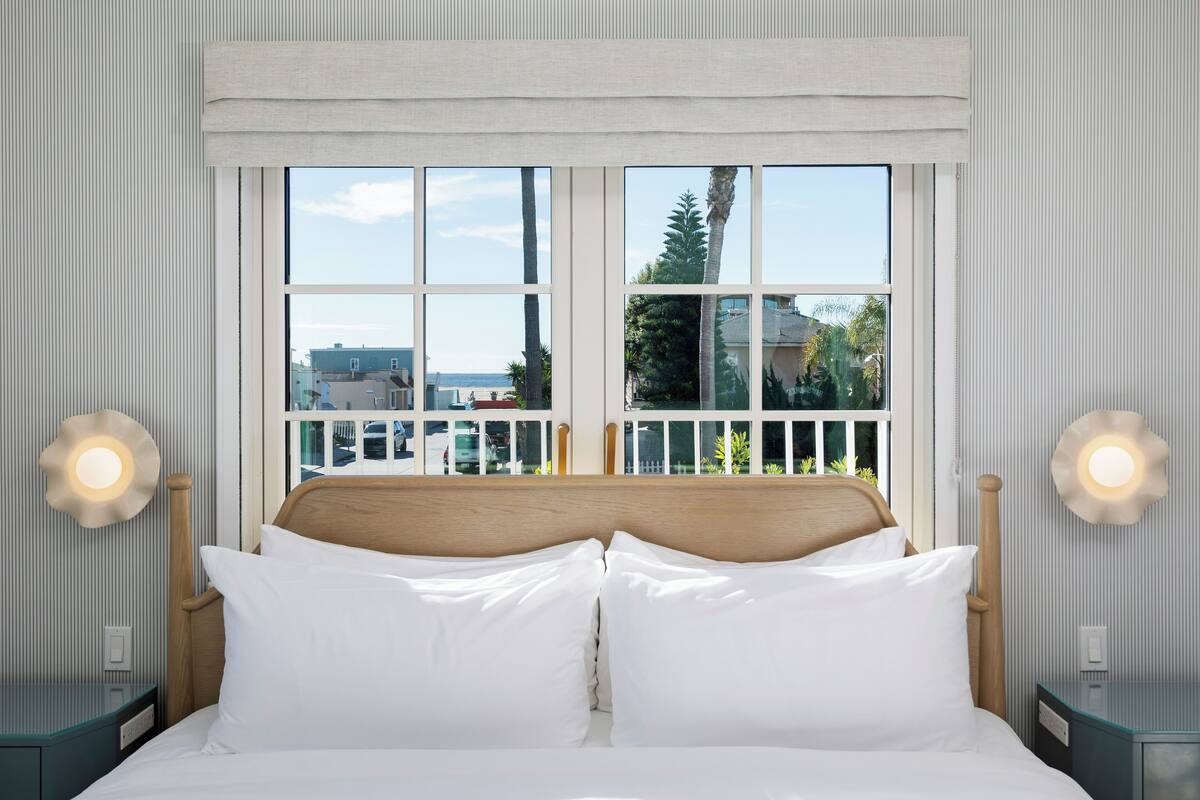 Ein ordentlich bezogenes Bett mit weißen Leinen, einem hölzernen Kopfteil und Blick durch ein großes Fenster auf Gebäude und Bäume.