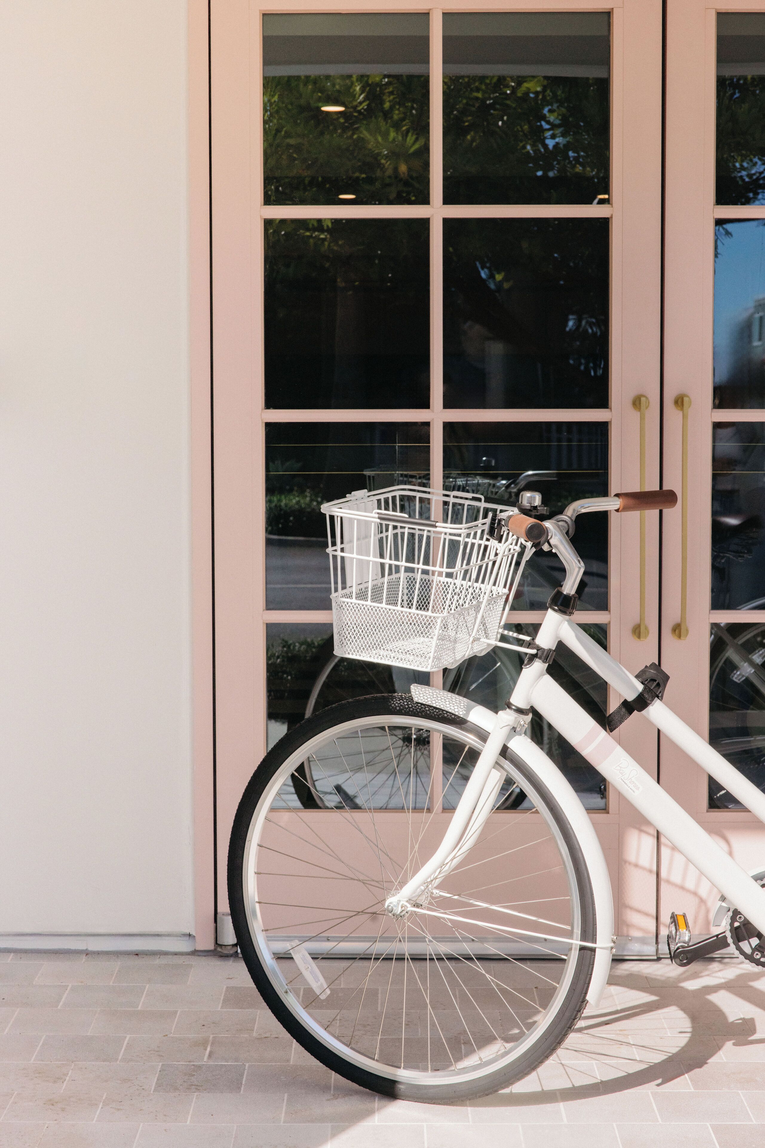 Una bicicleta blanca con canasta, estacionada frente a un edificio con ventanas amplias y una puerta.