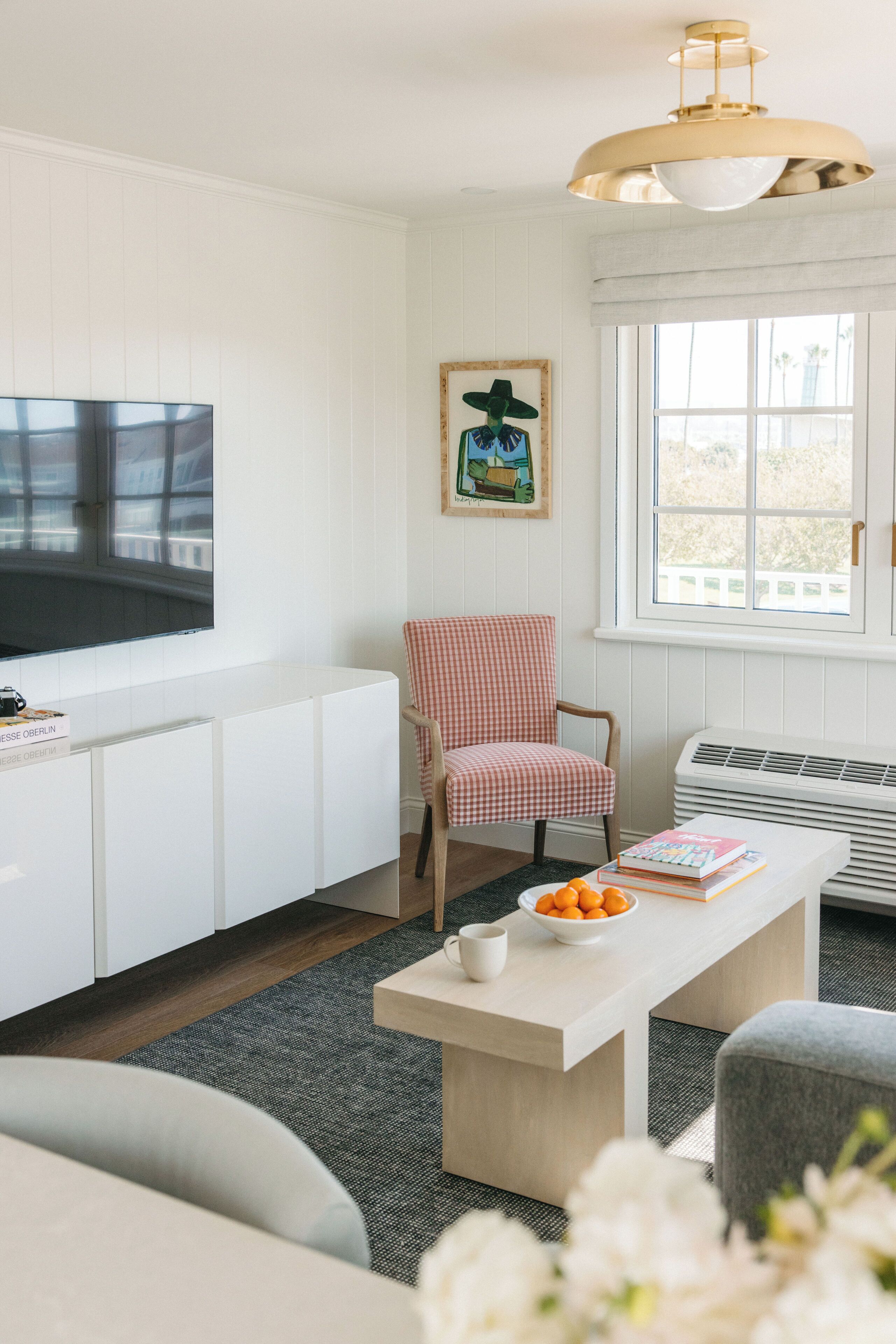 A living room with a flat-screen TV, a checkered armchair, a coffee table with books and oranges, and a window with a view.