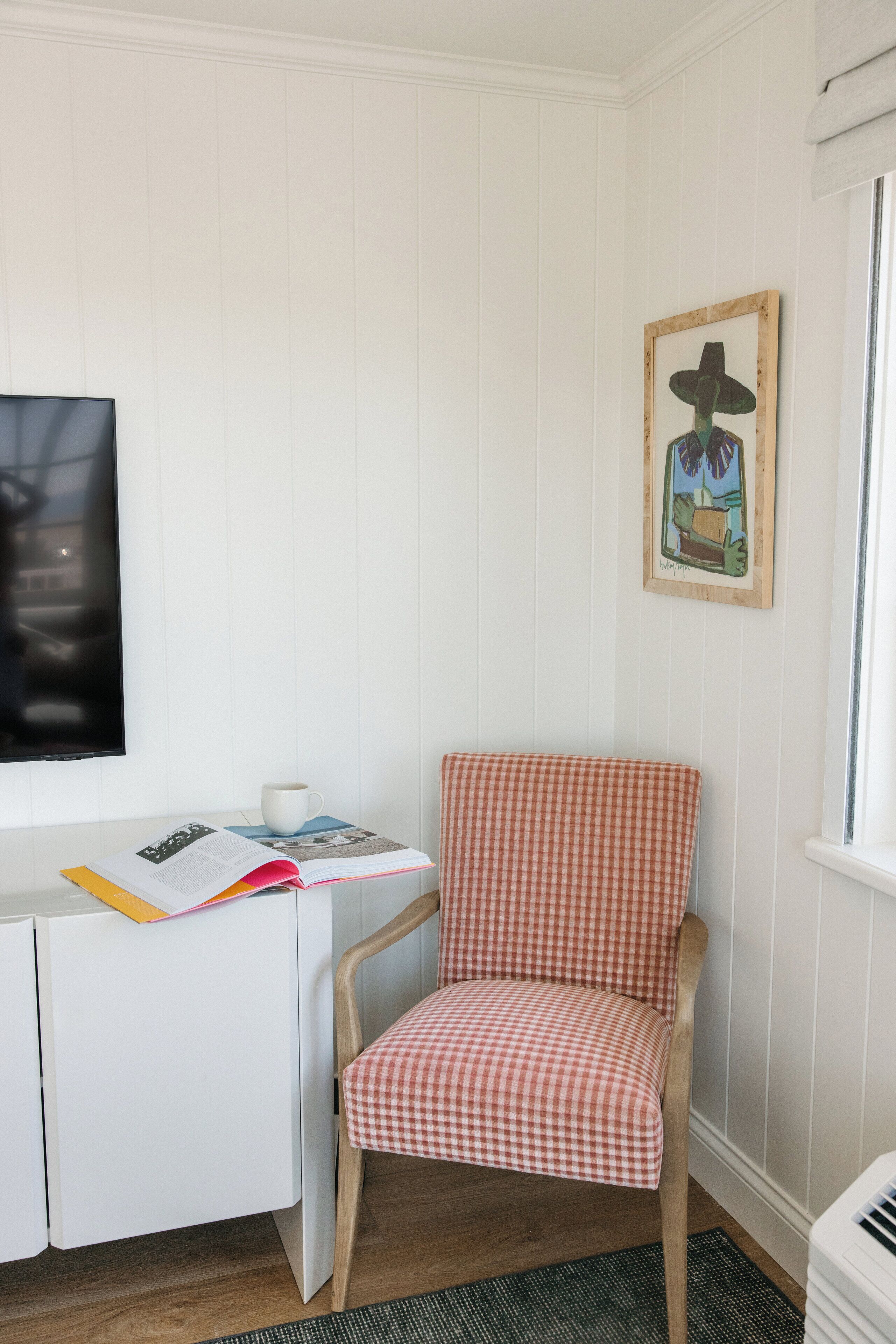 A corner room with a white desk, a checkered chair, a framed artwork, and a window.