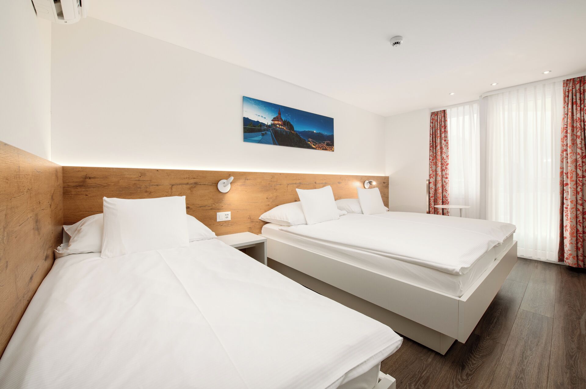 Een hotelkamer met twee bedden, een houten hoofdeinde, een nachtlampje en een schilderij van een stadsgezicht.