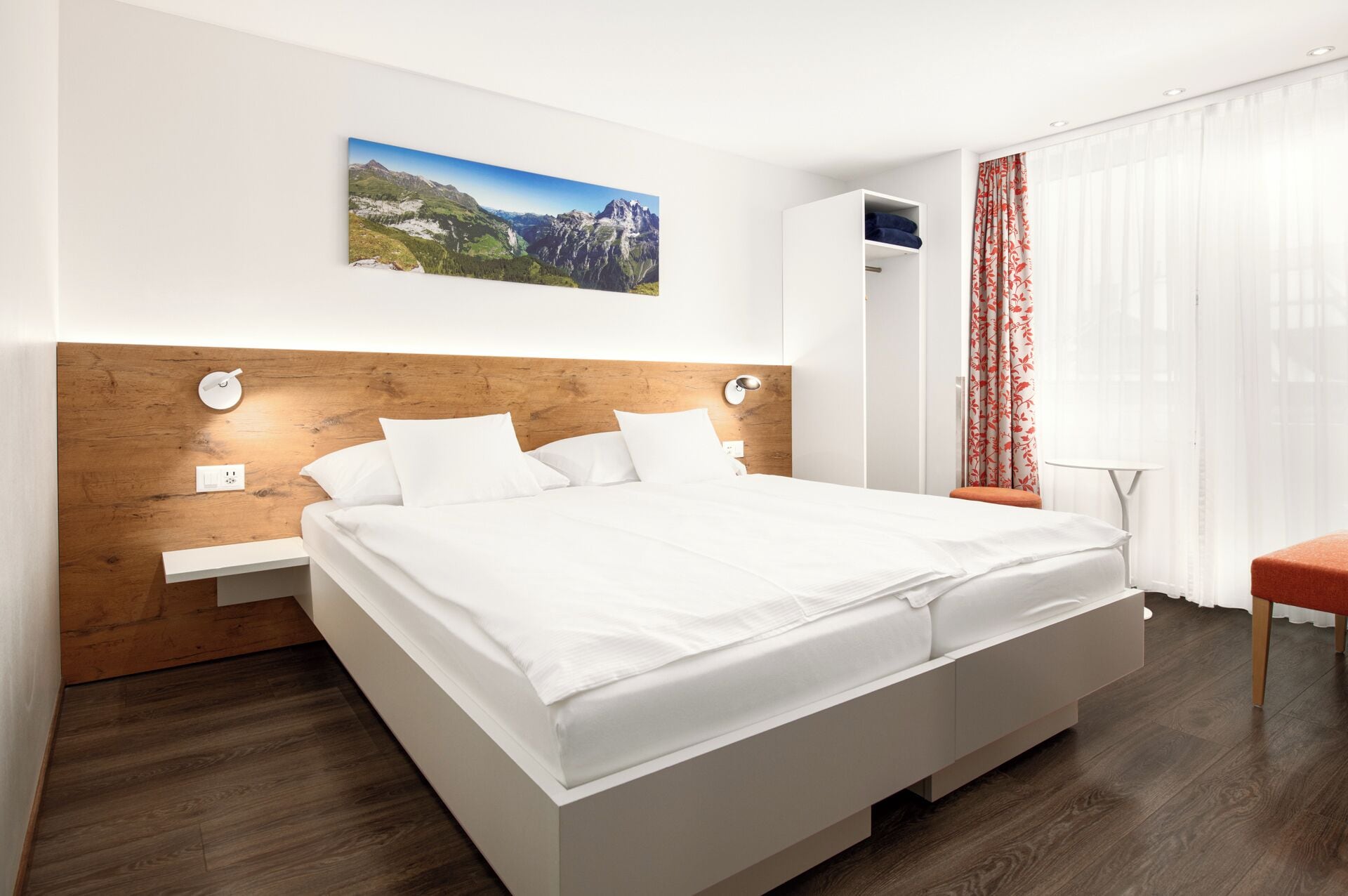 Une chambre d’hôtel moderne avec un grand lit, une tête de lit en bois, un tableau accroché au mur, une armoire et une fenêtre ornée de rideaux légers.