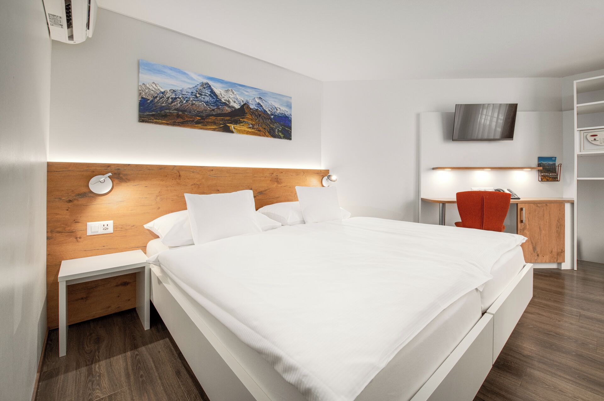 Ein modernes Hotelzimmer mit einem hölzernen Kopfteil, einem großen Bett, einem Schreibtisch mit Stuhl, einem Fernseher und einem Bergpanorama.