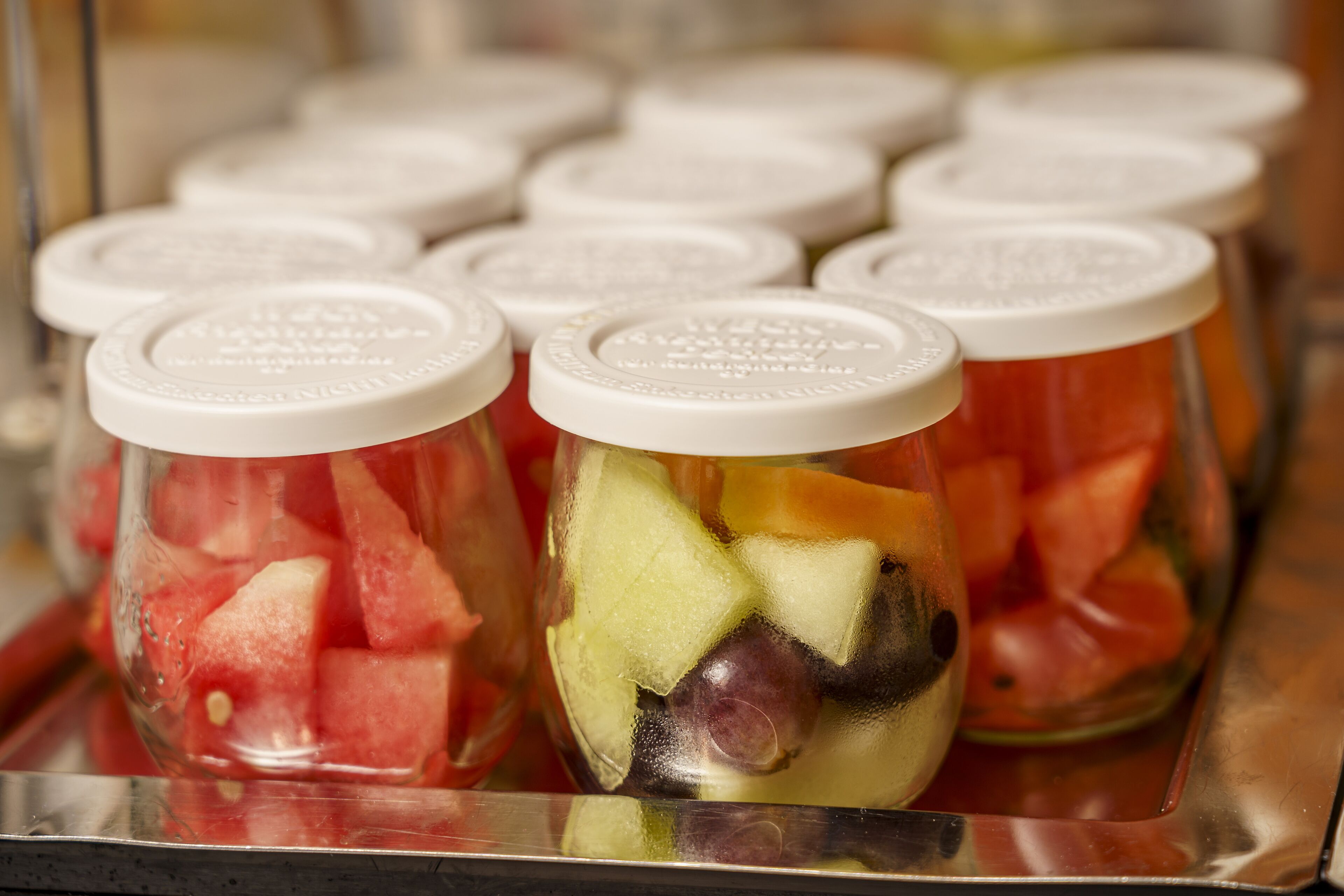 Varios frascos de vidrio con tapas blancas, llenos de diferentes frutas como sandía y melón.